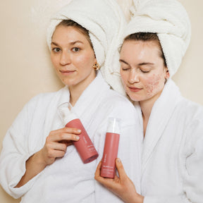 Zwei weiblich gelesene Personen in weißem Bademantel und weißem Handtuchturban halten jeweils eine Flasche clear skin in die Kamera. Eine Person schaut in die Kamera, die andere auf die Produkte